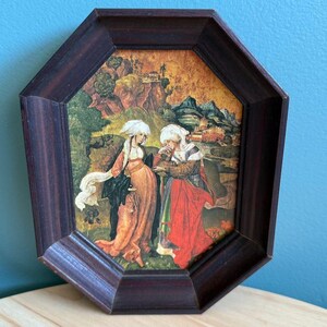 May include: A framed print of a painting depicting two women in medieval clothing standing in a landscape. The women are wearing long, flowing robes and head coverings. The background is a lush green forest with a distant mountain range.