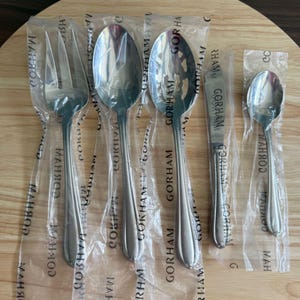 Può includere: Un set di cinque posate Gorham nuove e lucenti in acciaio inossidabile, tra cui una forchetta, due cucchiai, un cucchiaio forato e un coltello da burro, tutte in buste di plastica individuali, su una superficie di legno.