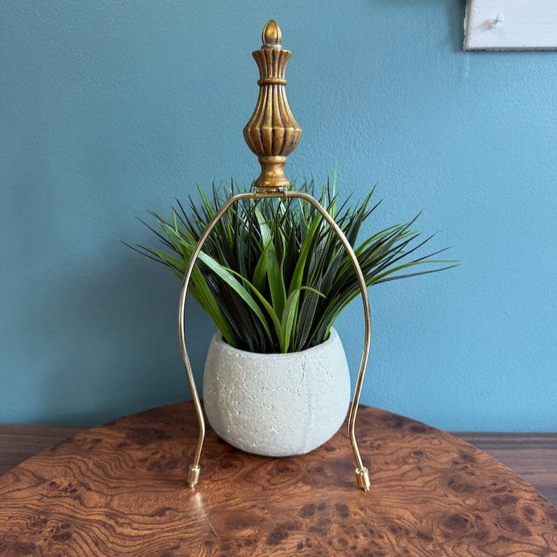 May include: A gold-colored lamp finial with a decorative design sits above a gold-colored lamp harp. The harp arches over a small, white, textured pot filled with green, spiky plants. The lamp base is not visible.