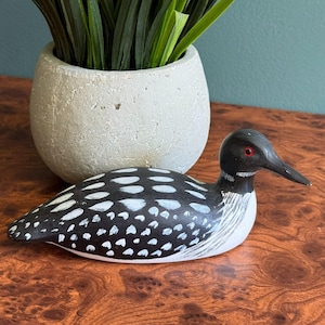 May include: A hand-painted wooden loon decoy featuring a black head with a red eye, a black back with white oval spots, and a white chest. The bird figure is positioned horizontally with its beak pointing to the right, showcasing detailed brushwork.