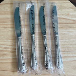 May include: Four new Gorham butter knives in clear plastic packaging. The stainless steel knives have a curved blade and a decorative handle. The word "GORHAM" is printed on the blade and handle.