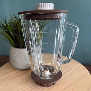 Op de afbeelding: Een heldere glazen blenderkan met een bruin deksel en basis. De kan heeft een handvat en maatstreepjes. Een wit en bruin deksel zit erop. De blender staat op een houten oppervlak.