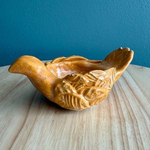 Puede incluir: Un plato de cerámica con forma de pájaro con un glaseado marrón dorado. El plato está decorado con detalles en forma de hojas y tiene un pico ligeramente curvado.