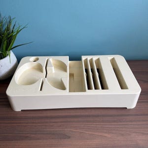 May include: Off-white plastic organizer with various compartments. The organizer has slots, curved sections, and a circular hole. The text "Cutter Cut" is visible on the side. The organizer is on a wooden surface with a blue background.