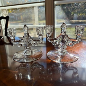 May include: Two clear glass candelabras with three candle holders each. The candelabras have a decorative, ornate design with a round base and a central, pointed finial. The glass reflects light, creating a sparkling effect. The candelabras are on a wooden surface.