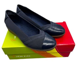 Puede incluir: Un par de bailarinas azul marino con punta redondeada y diseño cruzado. Los zapatos están sobre una caja colorida con las palabras "WEAR. EVER." impresas en el lateral.