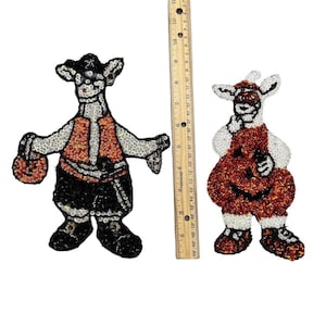 Könnte beinhalten: Zwei Halloween-Themenfiguren mit Pailletten. Eine Figur trägt einen schwarzen Hut, eine Weste und eine Hose und hält einen Kürbis. Die andere Figur trägt ein Kürbiskostüm. Die Figuren sind etwa 20 cm groß.