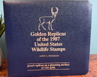 May include: A navy blue album titled "Golden Replicas of the 1987 United States Wildlife Stamps." The cover features a gold illustration of a deer and text in gold lettering. The text reads "proof replicas on a gleaming surface of 22kt gold."