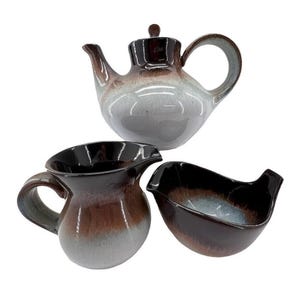 May include: A ceramic tea set with a teapot, creamer, and sugar bowl. The set features a gradient color scheme of brown, gray, and white. The teapot has a lid and a curved handle. The creamer has a handle and a spout. The sugar bowl is a small, open dish.