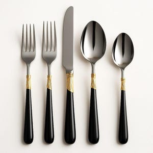 Puede incluir: Un juego de cubiertos de acero inoxidable de cinco piezas. El juego incluye dos tenedores, un cuchillo y dos cucharas. Cada pieza tiene un mango negro con un acento dorado cerca de la parte superior. Los utensilios están dispuestos sobre un fondo blanco.
