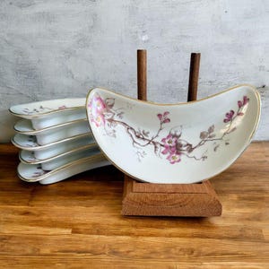 Puede incluir: Juego de seis fuentes de porcelana blanca con forma curva de barco. Cada fuente presenta un diseño floral con flores rosas y tallos marrones, acentuado con ribetes dorados. Las fuentes están apiladas sobre una superficie de madera.