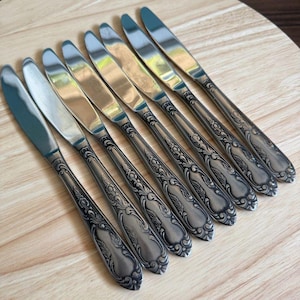 Puede incluir: Juego de ocho cuchillos de cena de plata con mangos ornamentados y detallados. Los cuchillos tienen un acabado pulido y están dispuestos en fila sobre una superficie de madera.