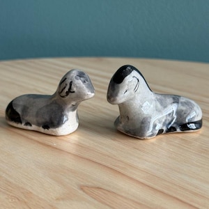Pode incluir: Duas pequenas estatuetas de cerâmica de ovelhas, uma deitada e outra sentada. Ambas são pintadas em tons de cinza e preto.
