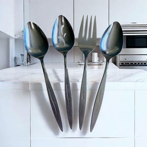 Puede incluir: Cuatro utensilios de cocina de acero inoxidable: dos cucharas, una cuchara ranurada y un tenedor. Las cucharas tienen forma de lágrima y el tenedor tiene cuatro púas. Dispuestos contra una encimera blanca en una cocina.