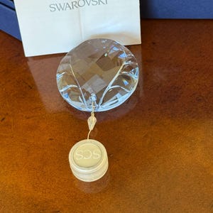 May include: A clear crystal ornament with a faceted design, hanging from a clear string. The ornament has a circular shape with a small, round tag that reads "SCS". The word "SWAROVSKI" is visible in the background.