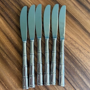 Könnte beinhalten: Set mit sechs Edelstahl-Buttermessern. Jedes Messer hat einen Bambus-Stil-Griff und eine gebogene Klinge. Die Messer sind auf einer Holzoberfläche angeordnet, was ihre polierte Oberfläche und ihr elegantes Design hervorhebt. Ideal zum Servieren von Butter oder Aufstrichen.