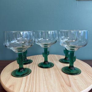 May include: Five clear margarita glasses with green cactus-shaped stems. The glasses have a rounded bowl and sit on a wooden serving board. The green stems add a unique, desert-inspired touch to the glassware.