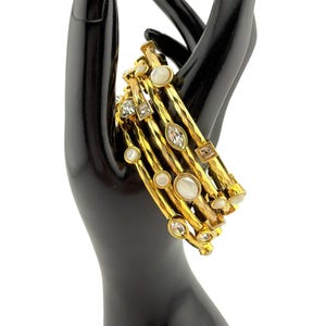 May include: A set of gold-tone bangles with various embellishments. The bangles feature clear, square, and oval-shaped crystals, along with round, white, pearl-like accents. The bracelets are displayed on a black mannequin hand.