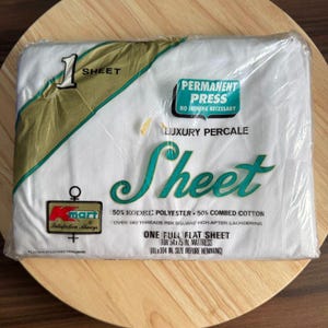 Könnte beinhalten: Ein verpacktes weißes Bettlaken mit dem Wort "Sheet" in türkisfarbener Schrift. Die Verpackung enthält den Text "Permanent Press" und "Luxury Percale". Das Laken ist für eine Matratze von 137x190 cm bestimmt.
