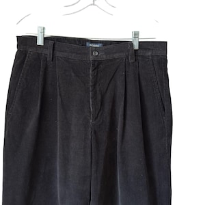 May include: Black corduroy trousers with a pleated front, hanging on a white hanger. The trousers have a button closure and pockets. The brand name "Dockers" is visible on the waistband.