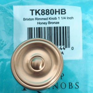 Puede incluir: Un pomo Brixton Honey Bronze, de forma circular, con un diámetro de aproximadamente 3,2 cm. El pomo tiene un botón central elevado y un borde. La etiqueta del producto dice "TK880HB" e incluye un código de barras.