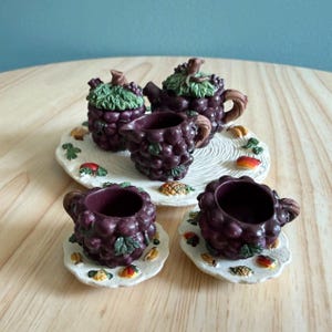Puede incluir: Un juego de té en miniatura que incluye una tetera, una jarra de leche y dos tazas con platillos, esculpidos como racimos de uvas de color morado oscuro con hojas verdes. Las piezas están dispuestas sobre una bandeja redonda con motivos frutales.