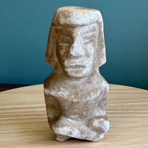 Puede incluir: Una escultura de piedra beige de una figura humana con una cabeza grande y rasgos estilizados. La escultura tiene un aspecto desgastado, con textura y detalles visibles. La figura está sentada con las manos sobre el regazo.