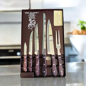 Può includere: Un set di coltelli Royal Hallmark in acciaio inossidabile in scatola con manici in legno scuro. Il set include un coltello da cucina, coltelli da bistecca, un coltello da intaglio, un coltello da pane seghettato e una forchetta da intaglio. La scatola ha il logo e il testo Royal Hallmark.