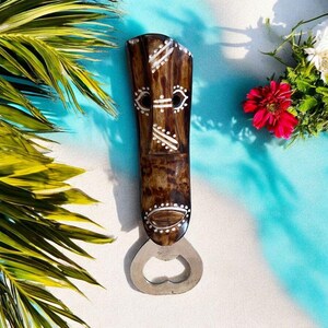 Può includere: Un apribottiglie in legno marrone con un design a maschera tribale. L'apribottiglie presenta dettagli dipinti di bianco e un apribottiglie in metallo argentato. Lo sfondo è un mix di blu e bianco con fogliame tropicale.
