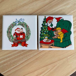 May include: Two square ceramic Christmas tiles. One tile features Santa Claus and a woman inside a wreath with a red bow. The other tile shows Santa Claus, a child, and a dog near a Christmas tree and a table with cookies.