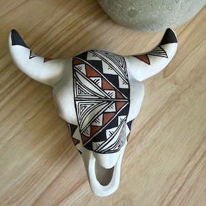 May include: A white ceramic bull skull wall decoration featuring a central geometric pattern in black and terracotta orange. The design includes repeating triangles and lines across the forehead and horns, with a hollow snout and pointed horns.