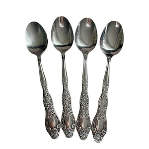 Puede incluir: Cuatro cucharas plateadas con mangos ornamentados con motivos florales. Las cucharas tienen cuencos ovalados y una superficie brillante y reflectante. Los mangos presentan volutas detalladas y una base decorativa y redondeada. Las cucharas están dispuestas en forma de abanico sobre un fondo blanco.