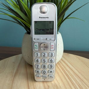 Pode incluir: Um telefone sem fio Panasonic prateado com um visor digital mostrando "Charge for 7h". O telefone tem um teclado com números e botões de função. É colocado em frente a uma planta verde em um vaso branco.