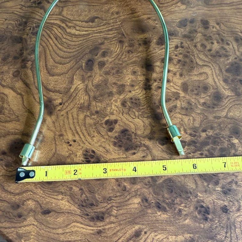 May include: A gold-colored lamp harp with a measurement of approximately 7 inches. The harp is curved and has a connector at each end. The background is a brown, wood-grain surface.