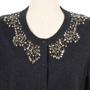 Puede incluir: Un cárdigan gris oscuro con un cuello decorativo con cuentas. El cuello presenta un intrincado diseño de cuentas transparentes y marrones. El suéter tiene una parte delantera abotonada y se muestra en un maniquí.