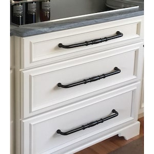 Peut inclure: Gros plan sur une armoire blanche à trois tiroirs, chacun équipé d'une poignée noire décorative. Les poignées ont un design incurvé avec une section centrale texturée. L'armoire fait partie d'une cuisine ou d'une salle de bain.