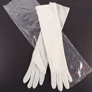 May include: A pair of long, pale grey gloves, possibly for formal occasions, are displayed on a dark surface. The gloves are made of a fine, textured fabric and extend well past the wrist. A clear plastic bag is partially visible in the background.