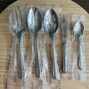May include: A set of five stainless steel Gorham serving utensils individually wrapped in clear plastic packaging. The set includes a three-tined fork, a large solid spoon, a large slotted spoon, a flat spreader, and a small spoon.
