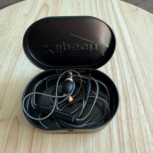 Puede incluir: Estuche ovalado negro abierto con el nombre de la marca "Kubecq" en relieve en la tapa. En el interior, auriculares con cable grises con detalles en negro y cobre enrollados. El estuche está sobre una superficie de madera.