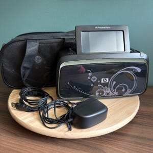 May include: A black HP Photosmart A646 compact photo printer with a flip-up screen, featuring a decorative swirl pattern on the front panel. Included are a black carrying case with a circular design and a black power adapter with a USB cable.