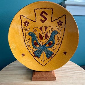 May include: A round, yellow decorative plate with a shield design featuring two blue birds, red floral accents, and a stylized symbol. The plate is displayed on a small wooden stand.