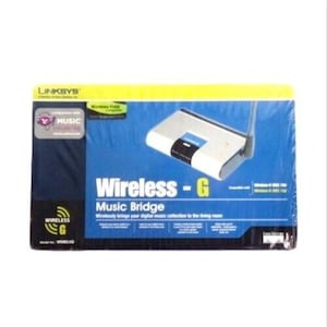 Può includere: Confezione originale di un dispositivo Linksys Wireless-G Music Bridge. La scatola è blu e gialla, con il nome del prodotto e una grafica del dispositivo. Il dispositivo è bianco e nero con un'antenna.
