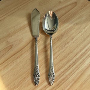 Puede incluir: Juego de cubiertos de plata con un cuchillo de mantequilla y una cuchara. El cuchillo tiene una hoja redondeada y la cuchara un cuenco ovalado. Ambos utensilios tienen detalles florales en los mangos. Sobre una superficie de madera marrón claro.