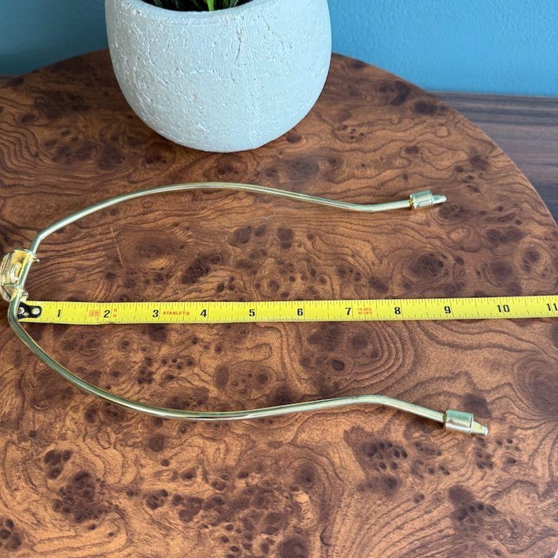 May include: A gold-colored lamp harp, approximately 10 inches long, rests on a wooden surface. The harp is curved and has a connector on each end. A small, light-colored pot with a plant is in the background.