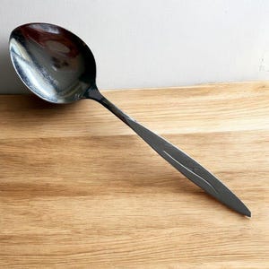 May include: A stainless steel serving spoon with a polished, reflective bowl and a dark, textured handle. The handle features a subtle, curved design. The spoon rests on a light brown wooden surface.