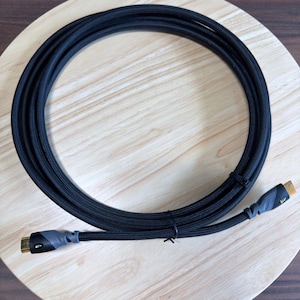 May include: A black braided cable coiled in a circular shape. The cable features two connectors with gold-colored tips and grey rubberized strain relief sections. The cord is secured with a thin black plastic tie to maintain its circular form.