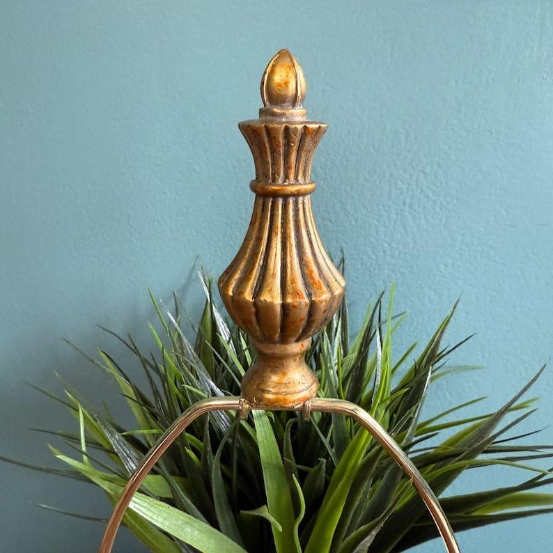 May include: A gold-colored, ornate finial with a ribbed design and a pointed top. The finial is part of a lamp, with a circular metal frame visible below. The background is a solid teal color, and the finial is set against a backdrop of green plants.