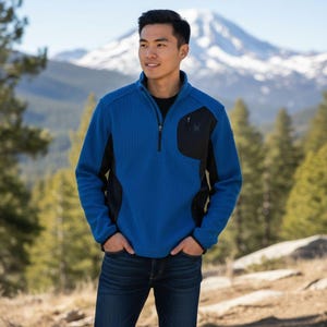 May include: A blue fleece jacket with black accents and a zippered chest pocket. The jacket has a black zipper and a small logo on the pocket. The person is wearing a black shirt underneath and blue jeans. The background features a mountain and trees.