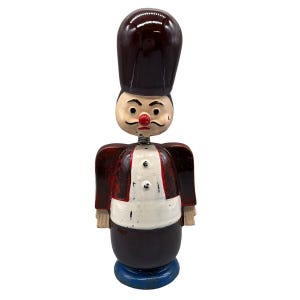 May include: A vintage wooden toy soldier with a tall, dark brown hat and a painted face. The soldier wears a red jacket with white accents and a blue base. The head is spring-mounted.