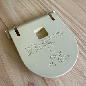 Peut inclure: Couvercle de blender en plastique blanc cassé avec le texte "FOR FOOD PROCESSOR OPERATION THIS COVER MUST BE POSITIONED OVER BLENDER WELL. PUSH TO OPEN." Le couvercle a une ouverture carrée et deux languettes.